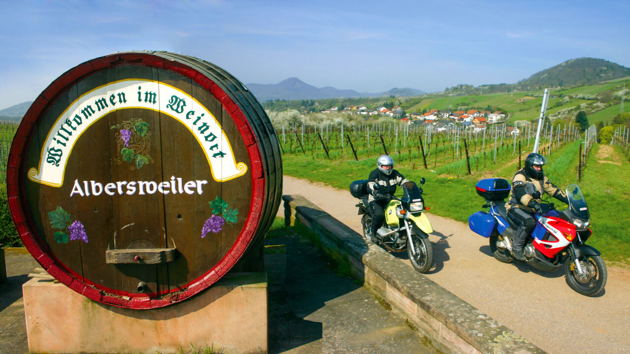 Großes Weinfass mit der Aufschrift Willkommen im Weinort Albersweiler, daneben zwei Motorräder vor Rebhängen