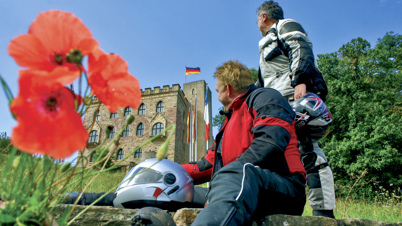Zwei Motorradfahrer rasten vor einem Schloss mit Fahne, im Vordergrund Mohnblumen